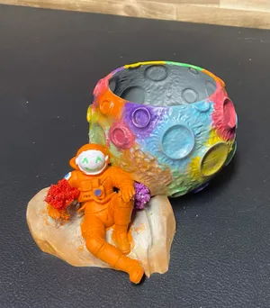 A rainbow-painted moon-shaped bowl with a small orange astronaut figure resting against it
