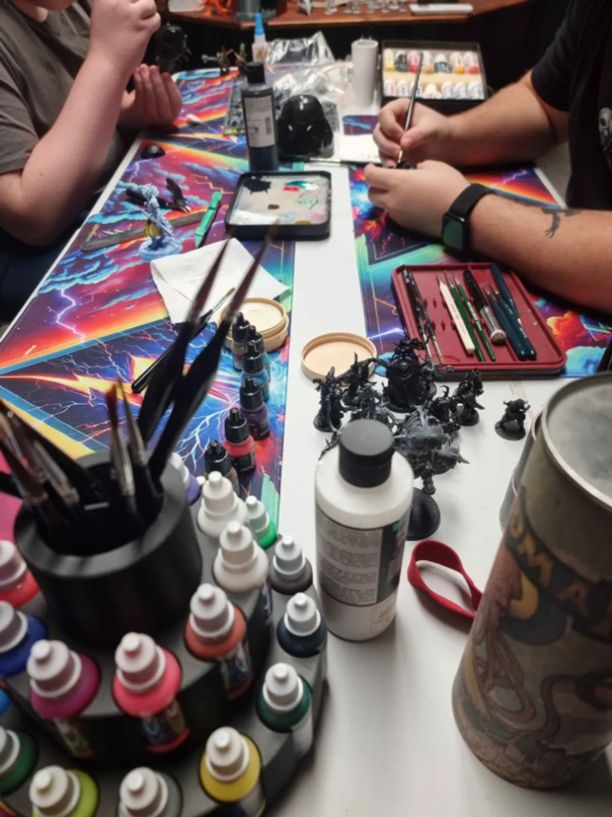 Two people painting miniatures across a shared workbench covered in paints, brushes and palettes