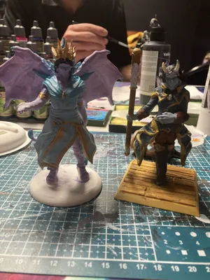 Two painted fantasy figures mid-session, one winged with a gold crown, one horned with a staff